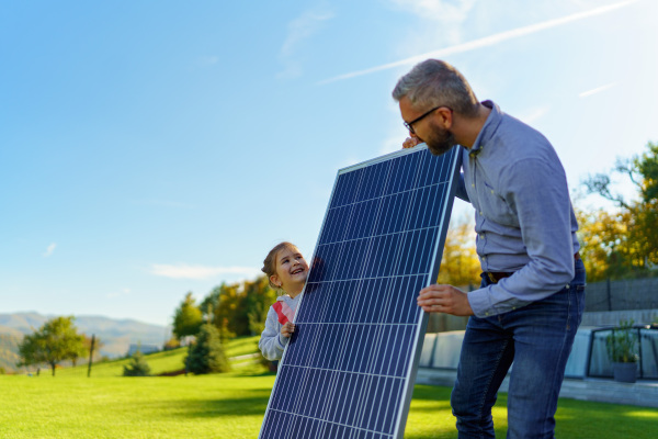 Father with his little daughter catching sun at solar panel,charging at the backyard. Alternative energy, saving resources and sustainable lifestyle concept.