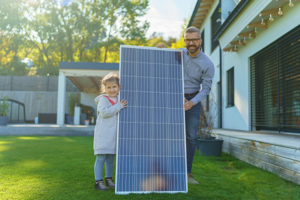 Father with his little daughter catching sun at solar panel,charging at the backyard. Alternative energy, saving resources and sustainable lifestyle concept.