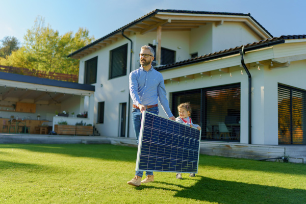 Father with his little daughter carring solar panel at the backyard. Alternative energy, saving resources and sustainable lifestyle concept.