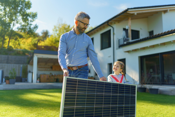 Father with his little daughter carring solar panel at the backyard. Alternative energy, saving resources and sustainable lifestyle concept.