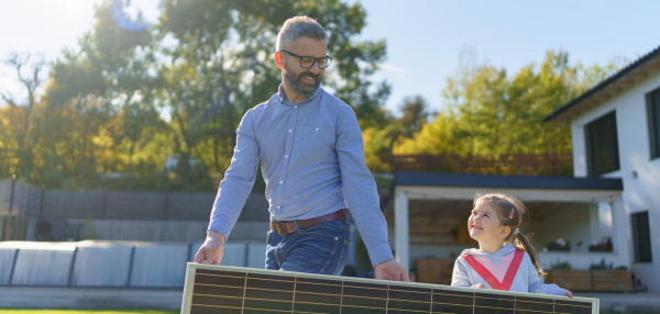 Father with his little daughter carring solar panel at the backyard. Alternative energy, saving resources and sustainable lifestyle concept.