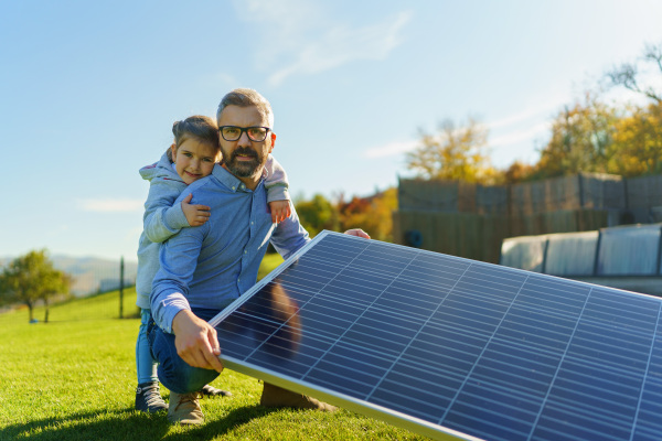 Father with his little daughter catching sun at solar panel,charging at the backyard. Alternative energy, saving resources and sustainable lifestyle concept.