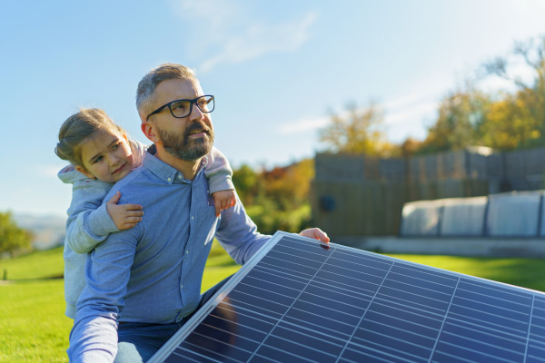 Father with his little daughter catching sun at solar panel,charging at the backyard. Alternative energy, saving resources and sustainable lifestyle concept.