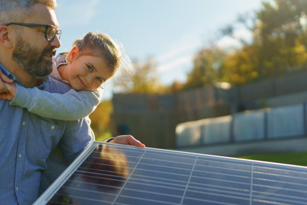 Father with his little daughter catching sun at solar panel,charging at the backyard. Alternative energy, saving resources and sustainable lifestyle concept.