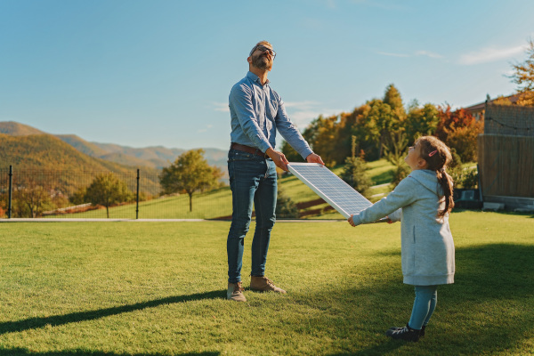 Father with his little daughter catching sun at solar panel,charging at the backyard. Alternative energy, saving resources and sustainable lifestyle concept.