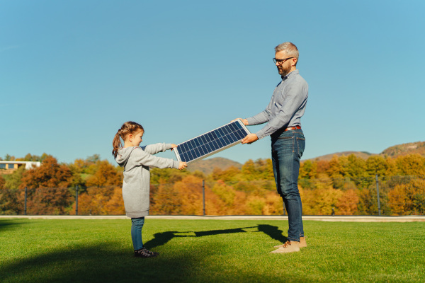 Father with his little daughter catching sun at solar panel,charging at the backyard. Alternative energy, saving resources and sustainable lifestyle concept.