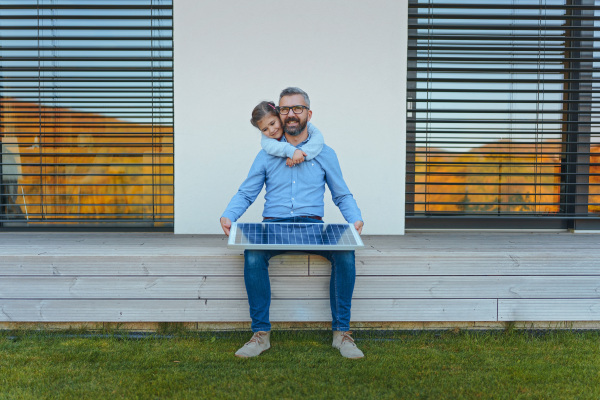 Father with his little daughter catching sun at solar panel,charging at the backyard. Alternative energy, saving resources and sustainable lifestyle concept.