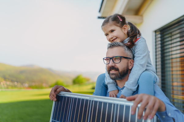 Father with his little daughter on piggyback, catching sun at solar panel,charging it at the backyard. Alternative energy, saving resources and sustainable lifestyle concept.