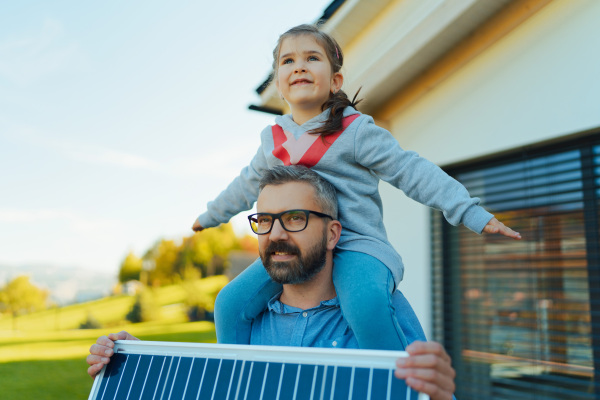 Father with his little daughter on piggyback, catching sun at solar panel,charging it at the backyard. Alternative energy, saving resources and sustainable lifestyle concept.