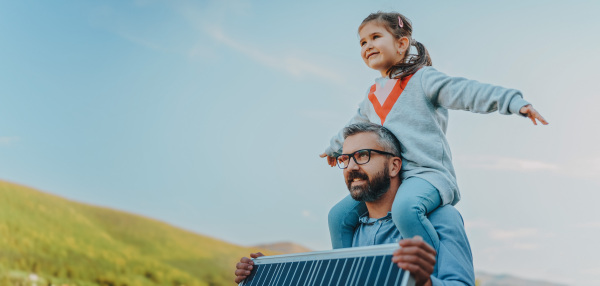 Father with his little daughter on piggyback, catching sun at solar panel,charging it at the backyard. Alternative energy, saving resources and sustainable lifestyle concept.