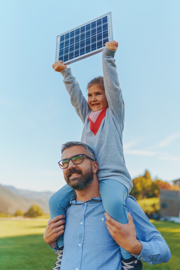 Father with his little daughter on piggyback, catching sun at solar panel,charging it at the backyard. Alternative energy, saving resources and sustainable lifestyle concept.