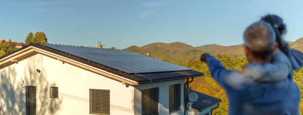 Rear view of dad holding her little girl in arms and showing at their house with solar panels.Alternative energy, saving resources and sustainable lifestyle concept.