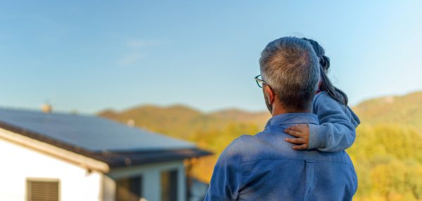 Rear view of dad holding her little girl in arms and looking at their house with solar panels.Alternative energy, saving resources and sustainable lifestyle concept.