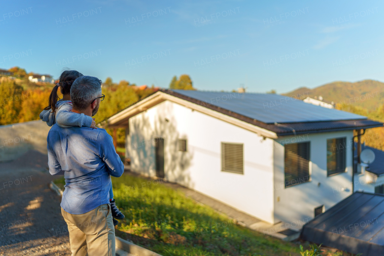 Rear view of dad holding her little girl in arms and looking at their house with solar panels.Alternative energy, saving resources and sustainable lifestyle concept.