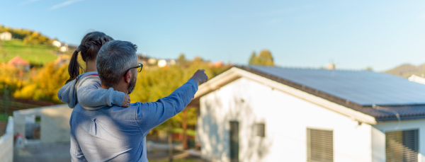 Rear view of dad holding her little girl in arms and showing at their house with solar panels.Alternative energy, saving resources and sustainable lifestyle concept.