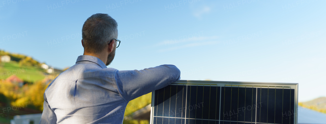 Rear view of businessman holding solar panel, standing outdoor at a garden.