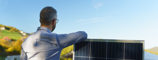 Rear view of businessman holding solar panel, standing outdoor at a garden.