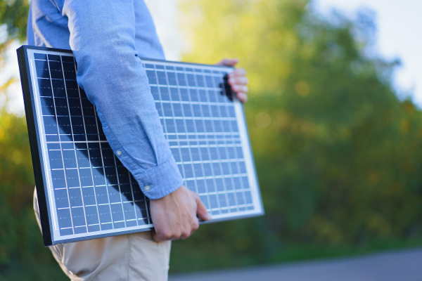 Mid section of businessman holding solar panel, standing outdoor at a garden.