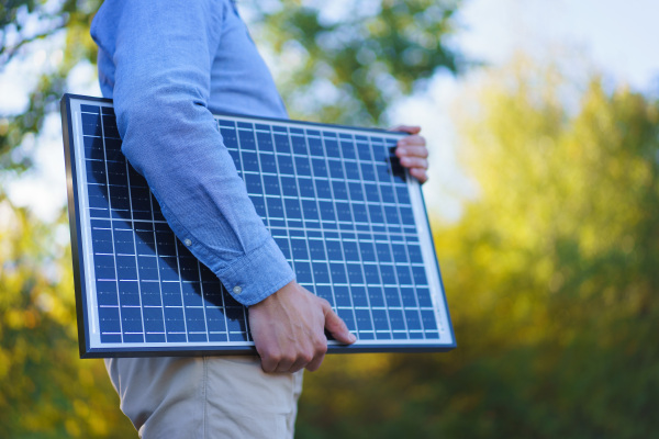 Mid section of businessman holding solar panel, standing outdoor at a garden.