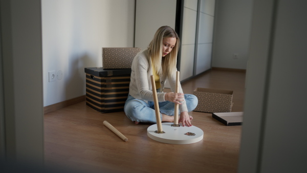 A single woman is assembling furniture in his first own apartment, putting together parts of wooden chair. The young woman living a single life, enjoying his own space.