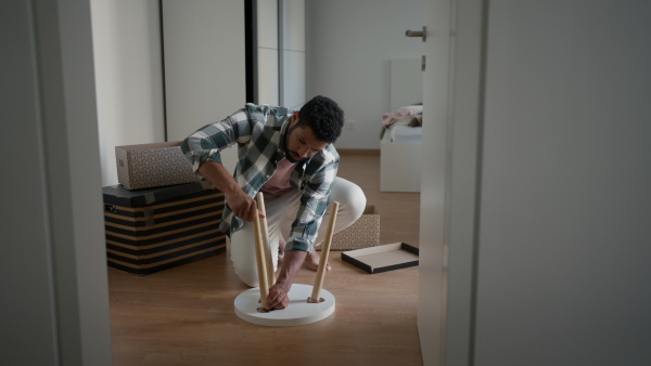 A single man is assembling furniture in his first own apartment, putting together parts of wooden chair. The young man living a single life, enjoying his own space.