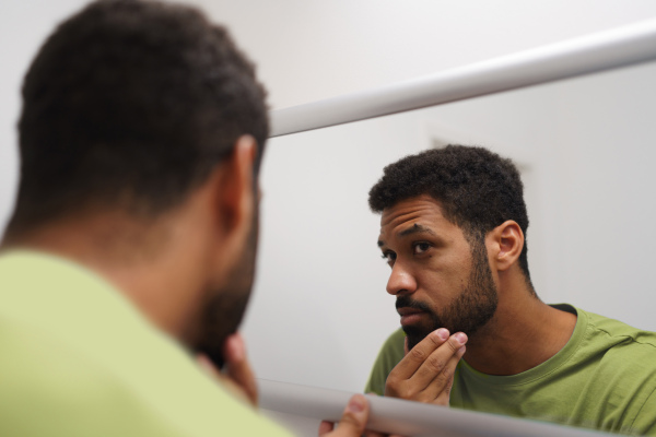 Handsome man looking at himself in the mirror, feeling anxious and depressed in the morning. Skincare routine for young man.