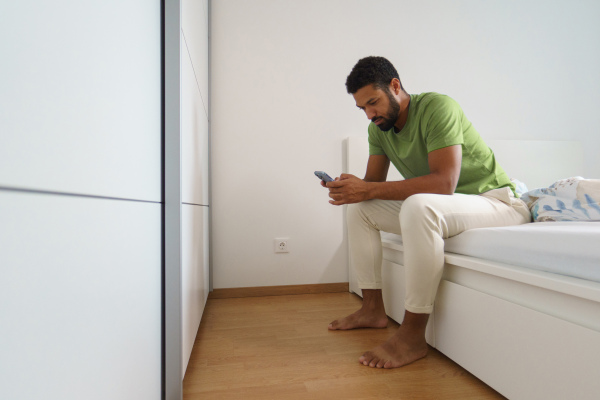 Single man waking up alone in his bed. Sitting on bed and looking at smartphone, reading text messages.