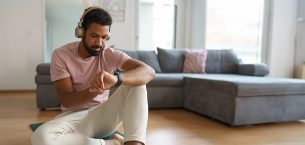 Young man resting after home workout, checking his exercise performance on smartwatch. Concept of morning or evening workout routine, listening to music through wireless headphones