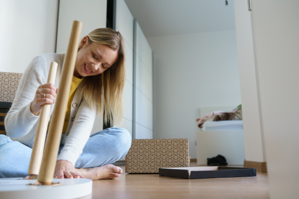 Young woman assembling furniture in her first own apartment, putting together parts of wooden chair. Single woman living a single life, enjoying own space.