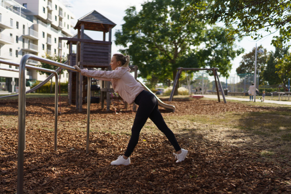 Woman exercises outdoors at an urban outdoor gym, stretching. Exercising after work for good mental health, physical health, and relieving stress and boost mood.