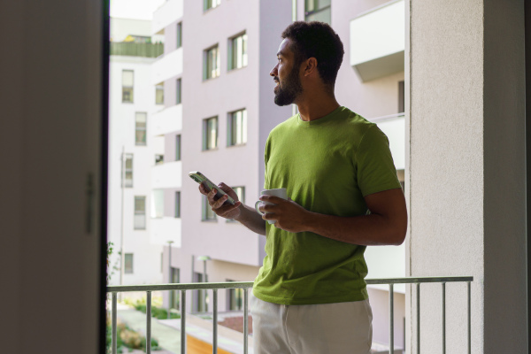 Single man waking up, drinking cup of morning coffee, looking out of window, balcony. Morning or weekend routine for single man.