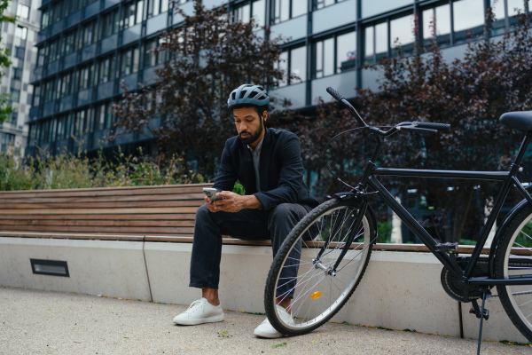 City commuter, sitting on bench, scrolling on smartphone. Businessman traveling from work to home on bike. City lifestyle, full lenght shot with copyspace.