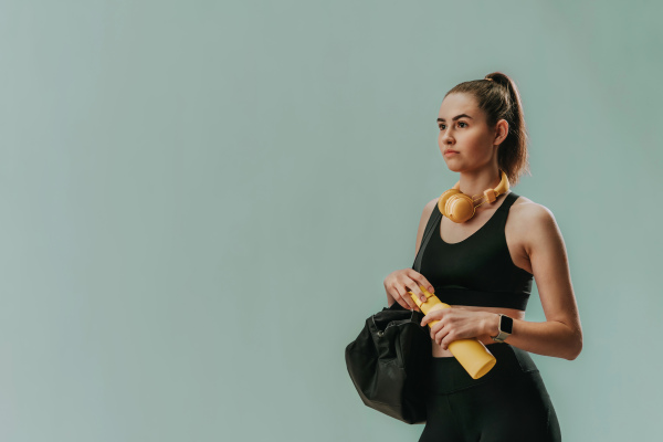 On the way to gym. A young sporty woman in sportswear with fitness bag, wireless headset and bottle over green background, copy space