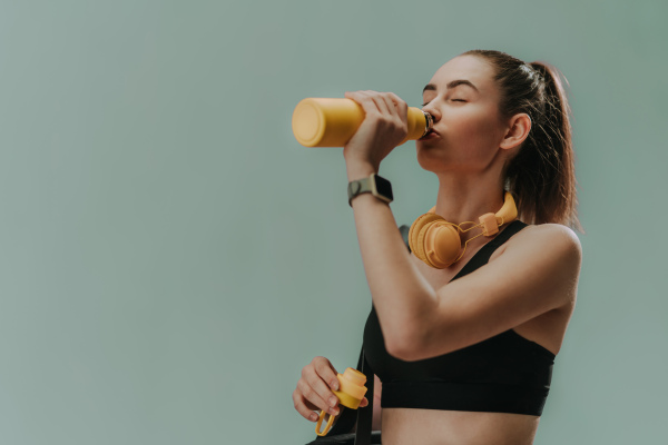 A young sporty woman in sportswear with wireless headset drinking from bottle over green background, copy space