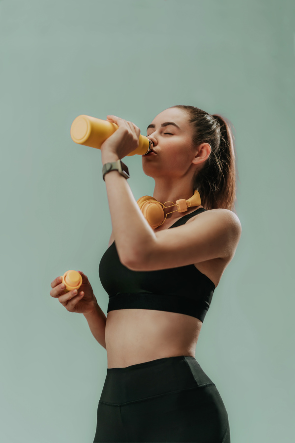 A young sporty woman in sportswear with wireless headset drinking from bottle over green background, copy space