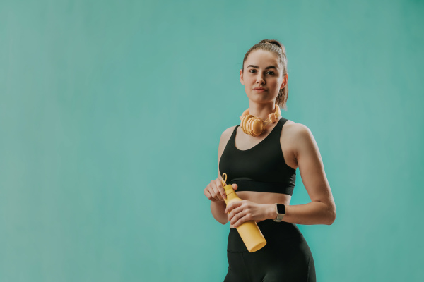 A young sporty woman in sportswear with wireless headset and bottle over green background looking at camera.