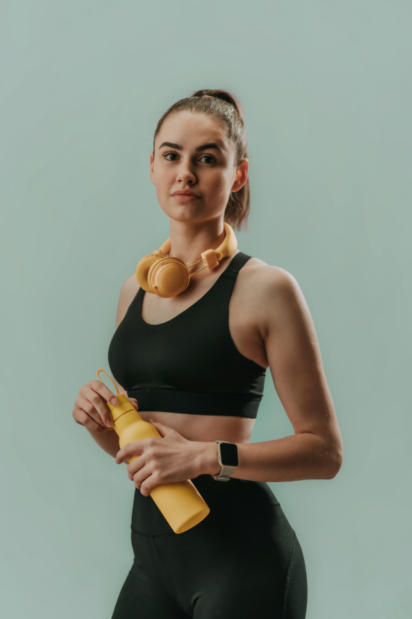 A young sporty woman in sportswear with wireless headset and bottle over green background looking at camera.