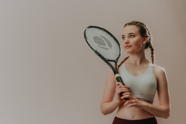 A young sporty woman in sports clothes holding tennis racket on her shoulder while standing against beige background, copy space