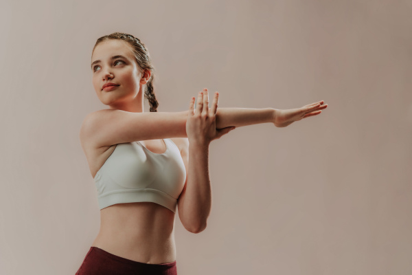 A young sporty woman in sports clothes stretching arms against beige background, copy space