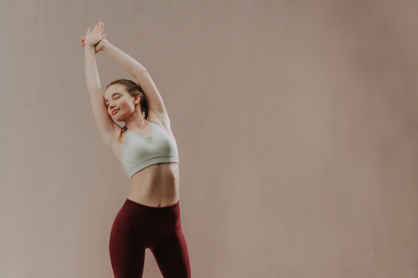 A young sporty woman in sports clothes stretching arms against beige background, copy space