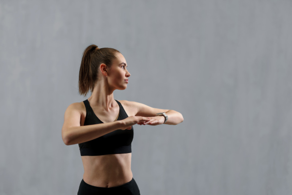 A young sporty woman in sports clothes stretching arms against grey background, copy space