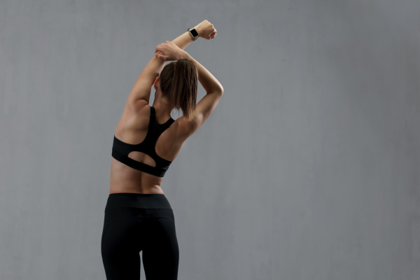 A rear view of young sporty woman in sports clothes stretching arms against grey background, copy space