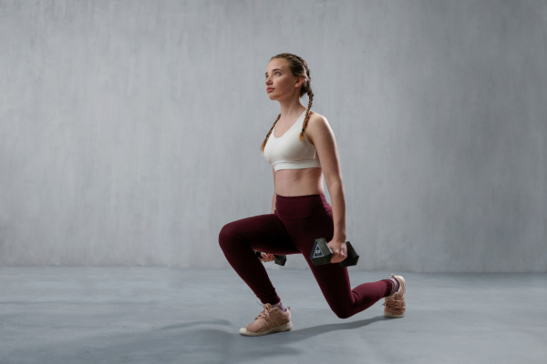 A young athletic woman in sportswear doing squat using dumbbells, posing in studio with grey background.