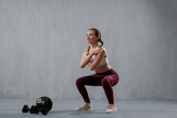 A young athletic woman in sportswear doing squat using dumbbells, posing in studio with grey background.