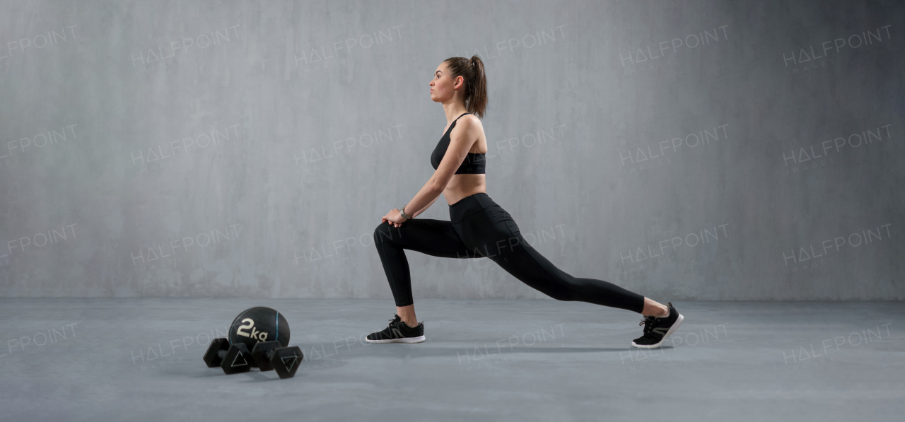 A young athletic woman in sportswear doing squat using dumbbells, posing in studio with grey background.