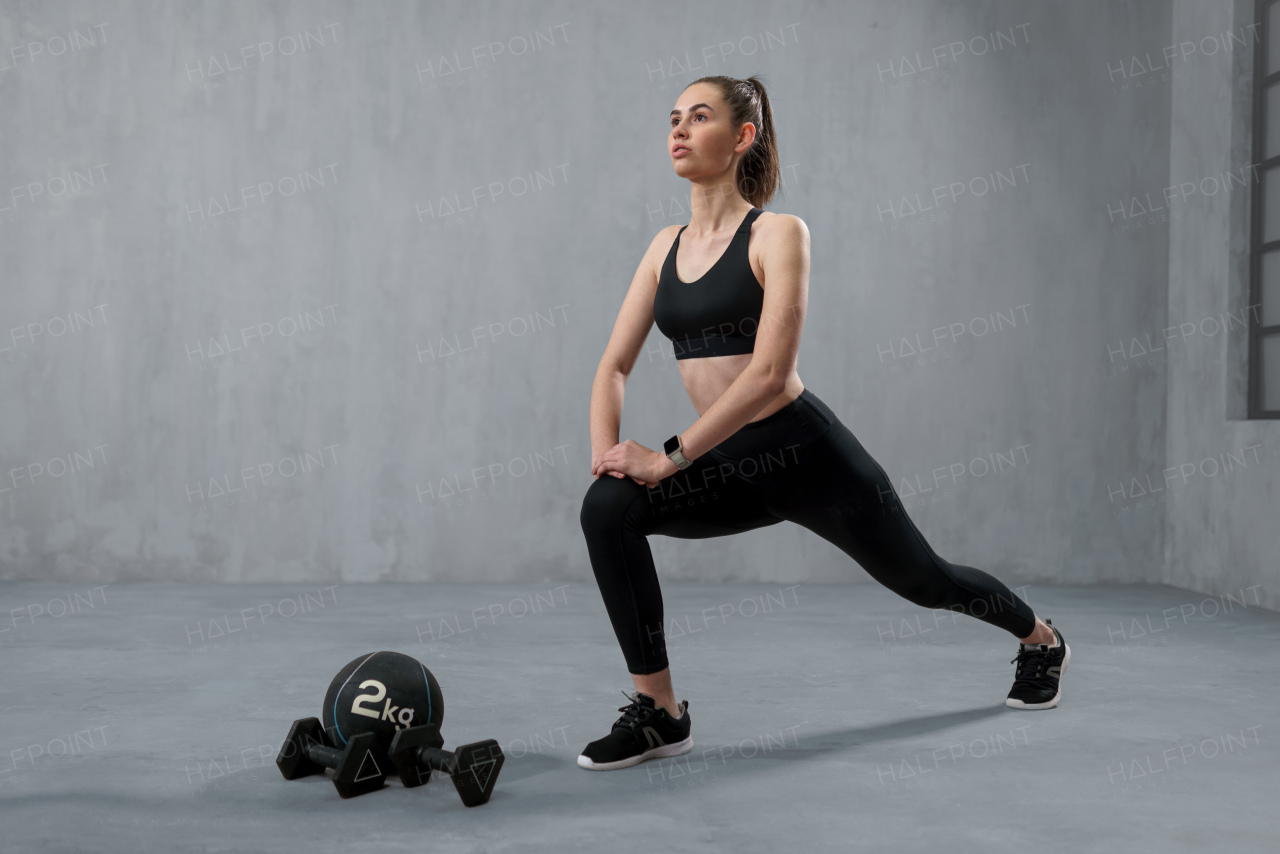 A young athletic woman in sportswear doing squat using dumbbells, posing in studio with grey background.