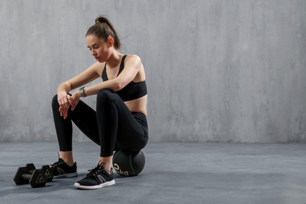 A tired young sporty woman having rest after workout, sitting on floor at gym , over grey background.