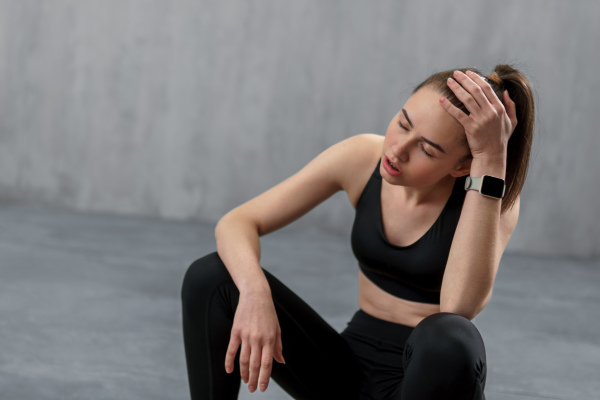 A tired young sporty woman having rest after workout, sitting on floor at gym , over grey background.