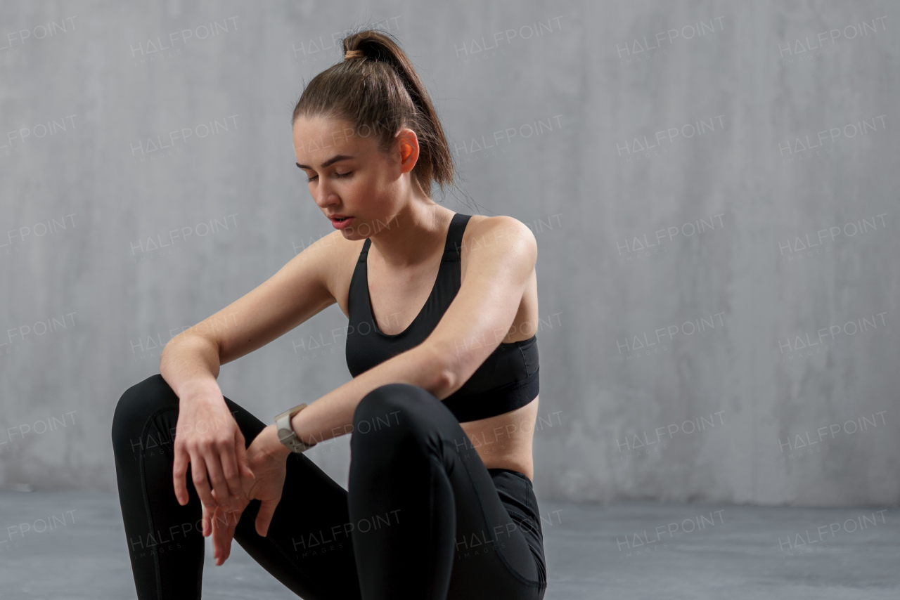 A tired young sporty woman having rest after workout, sitting on floor at gym , over grey background.