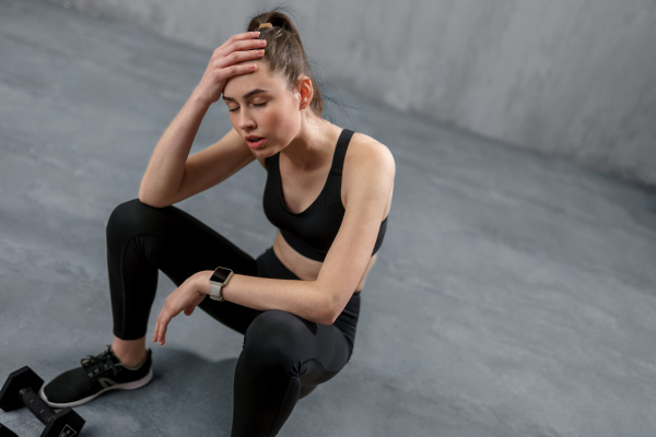 A tired young sporty woman having rest after workout, sitting on floor at gym , over grey background.